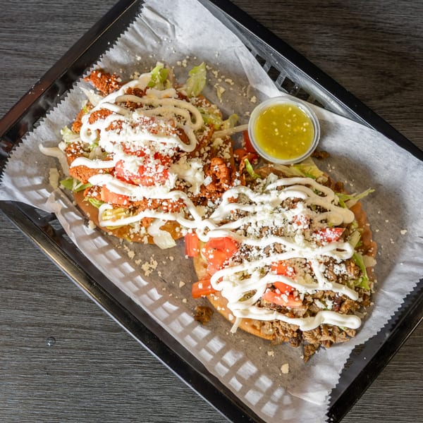 Two Mexican sopes with meat, lettuce, tomatoes, cheese, and crema, with a side of green salsa.