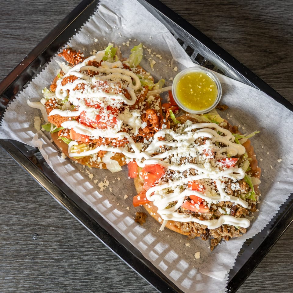 Two Mexican sopes with meat, lettuce, tomatoes, cheese, and crema, with a side of green salsa.