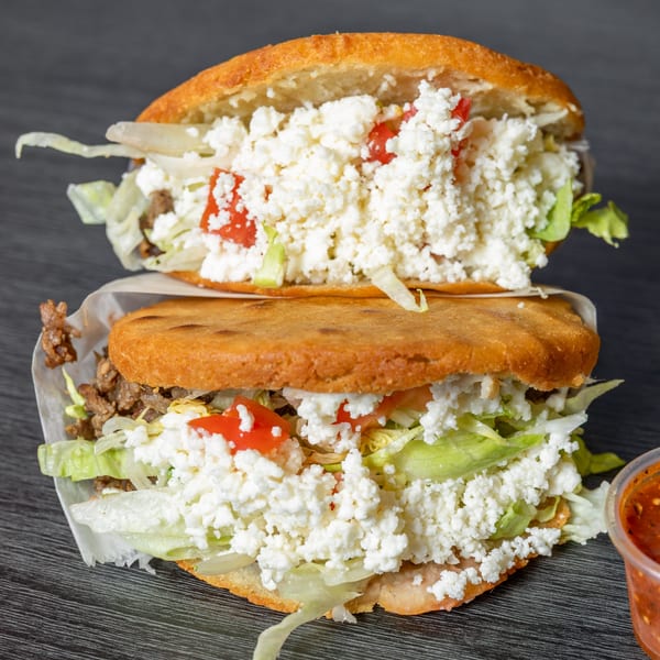 Two Tacos Ponchos gorditas, stacked, filled with meat, lettuce, tomatoes, and crumbled white cheese.