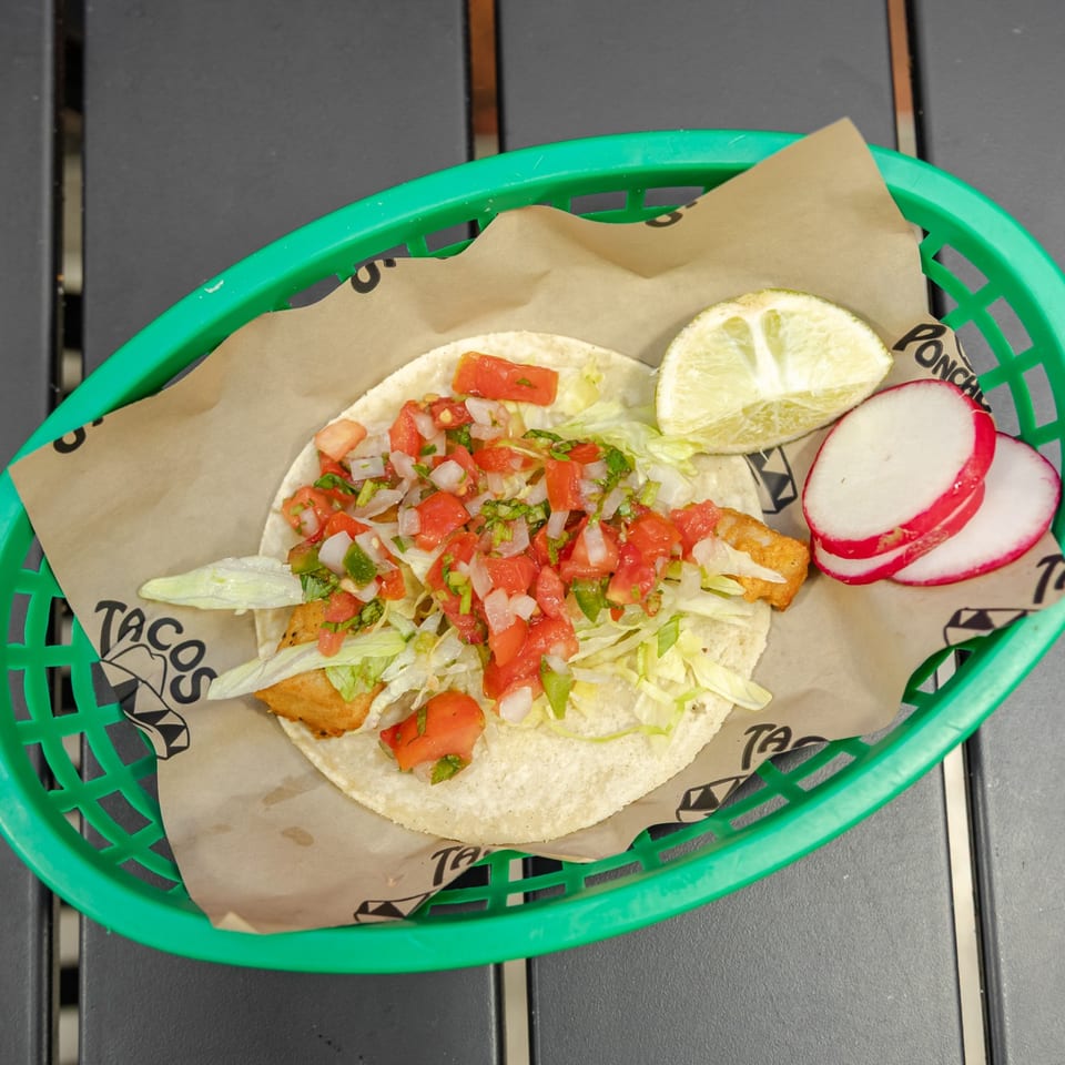 Tacos topped with shredded lettuce and pico de gallo, served with lime and sliced radishes in a green basket.