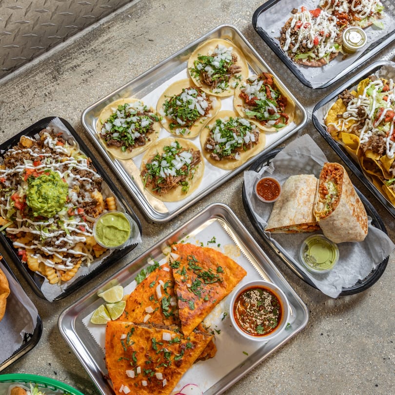 An overhead shot of various Mexican dishes, including tacos, nachos, burritos, and quesadillas, on a concrete surface.