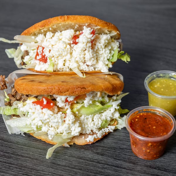Two arepas filled with meat, lettuce, tomatoes, and crumbled white cheese, with two salsa cups.