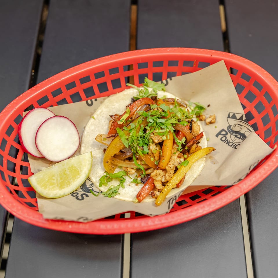 Two soft tacos filled with grilled chicken, peppers, onions, and cilantro in a red basket with radishes and lime.