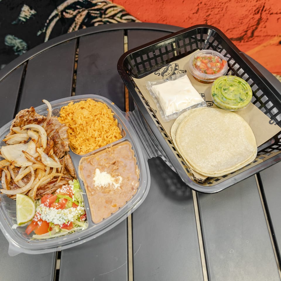 Mexican takeout meal with grilled meat, rice, beans, salad, tortillas, salsa, guacamole, and lime on an outdoor table.