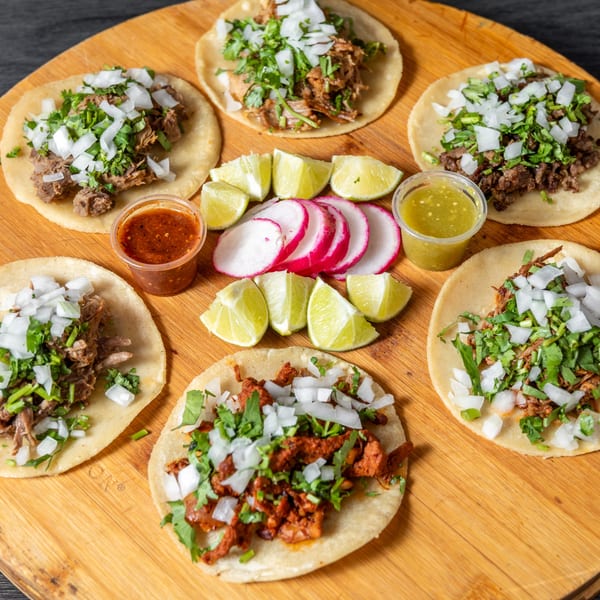 Six Tacos Ponchos tacos with various meats, cilantro, onions, lime, radishes, and salsas on a wooden board.