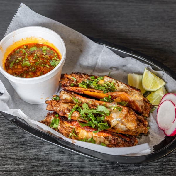 Four birria tacos with melted cheese and cilantro, a cup of consommé, lime wedges, and radish slices.