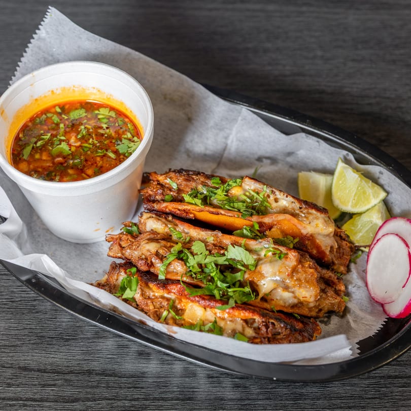 Four birria tacos with melted cheese and cilantro, a cup of consommé, lime wedges, and radish slices.