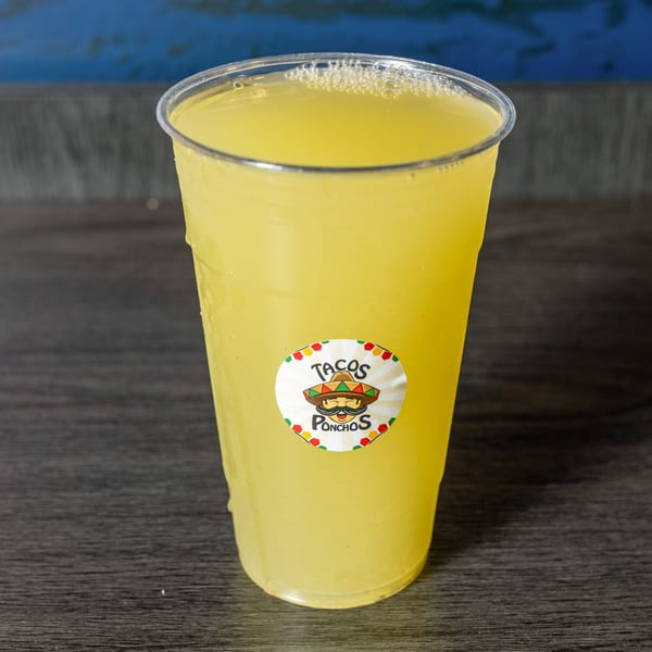 A clear plastic cup of yellow lemonade with a Tacos Ponchos logo sticker on a dark wooden surface.