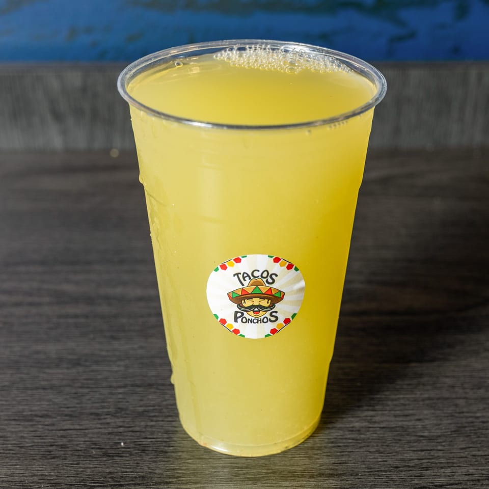 A clear plastic cup of yellow lemonade with a Tacos Ponchos logo sticker on a dark wooden surface.