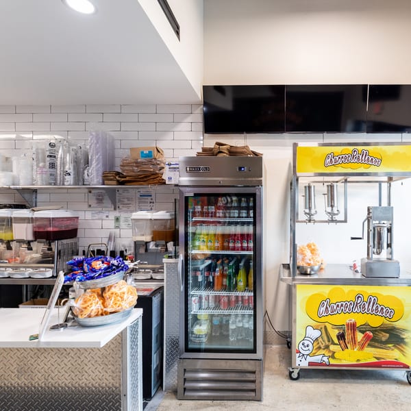 A Tacos Ponchos kitchen area with a drink fridge, churro machine, and food prep counter.