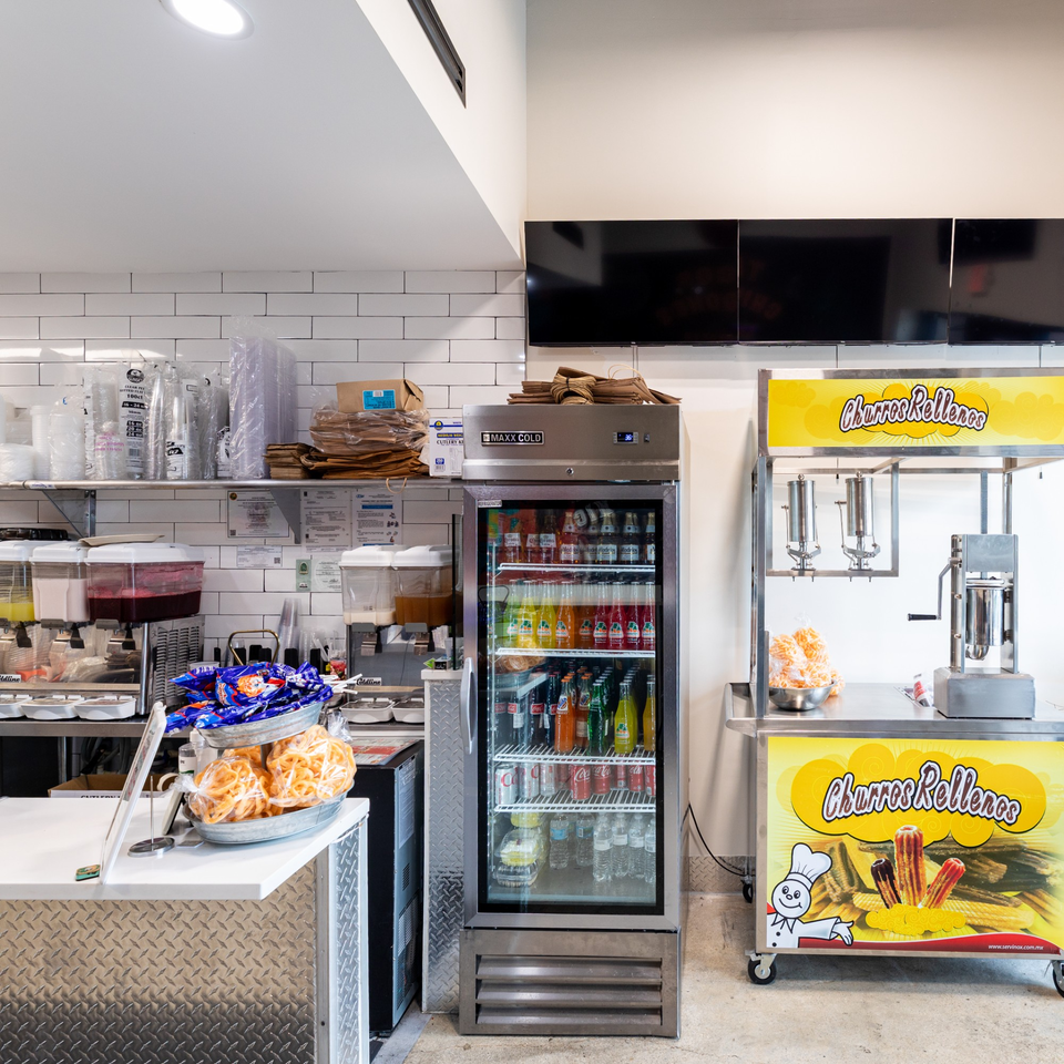 A Tacos Ponchos kitchen area with a drink fridge, churro machine, and food prep counter.