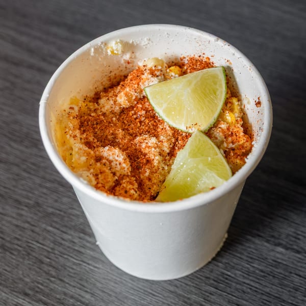Mexican street corn (Esquites) in a white cup, topped with chili powder, cheese, and lime wedges.