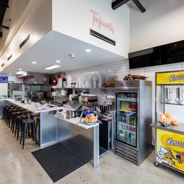 Interior of Tacos Ponchos showing a long counter with stools, kitchen area, and churro cart.