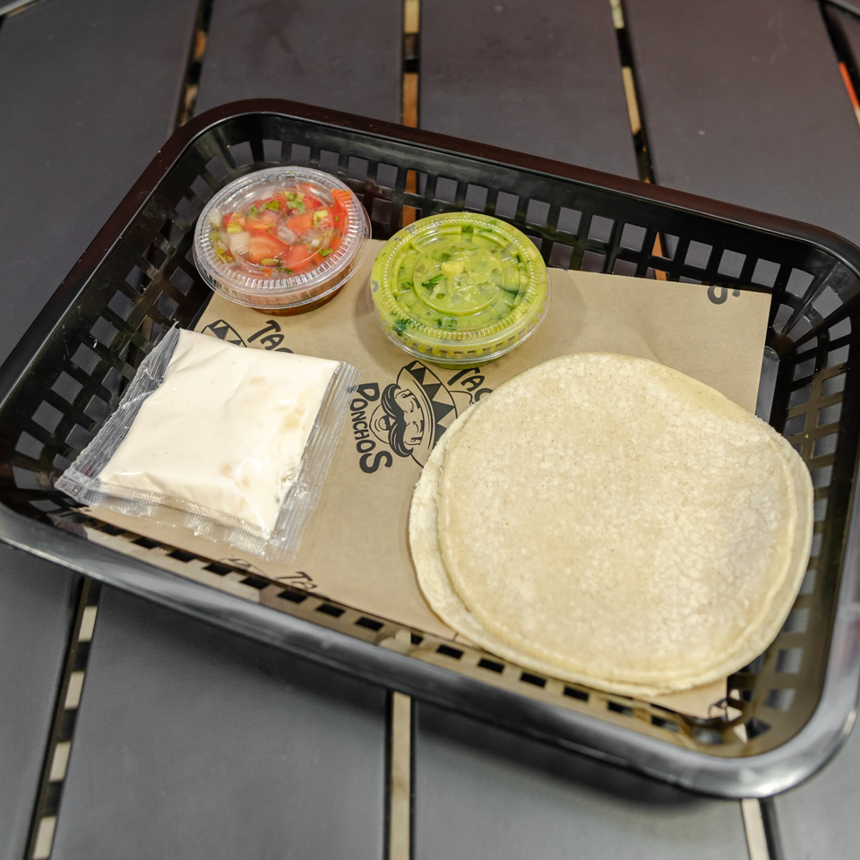 Basket holding corn tortillas, a creamy sauce packet, guacamole, and fresh salsa on branded restaurant paper.